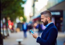 Hipster manager holding smartphone, texting outside in the street