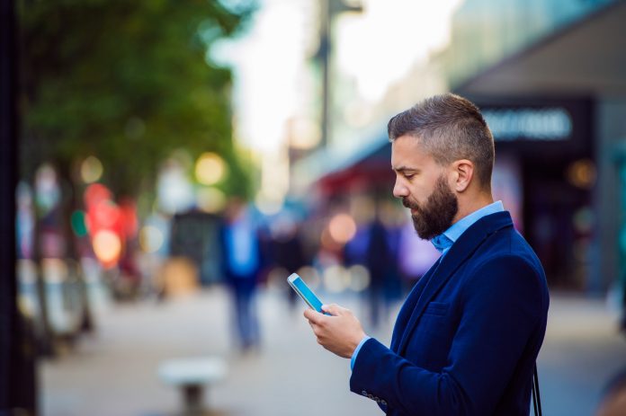 Hipster manager holding smartphone, texting outside in the street